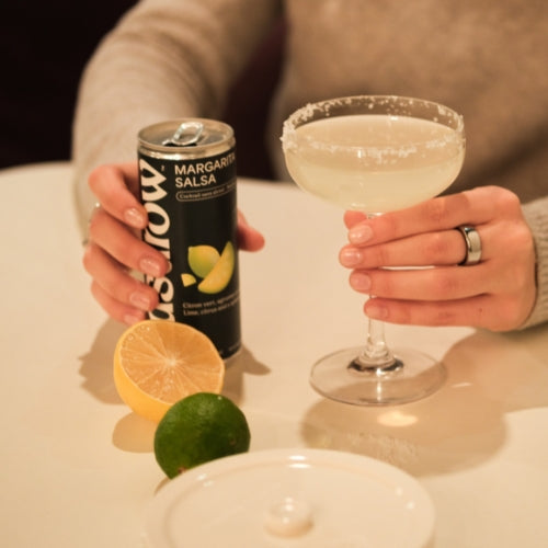 Person holding a Margarita Salsa mocktail and a can, with fresh limes and lemons on a table.